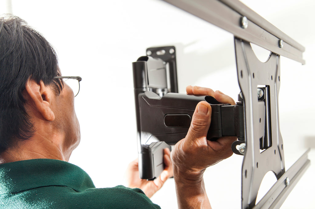 A man with black hair and glasses holds a metal tv wall mount by the arm so the bracket and adapter plate are visible.