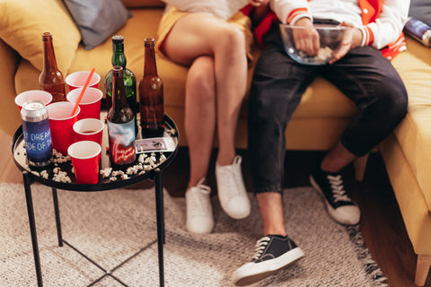 Two people blurred sitting down on a yellow couch in the background with a small circular table in the foreground filled with beer bottles, popcorn, and plastic cups.