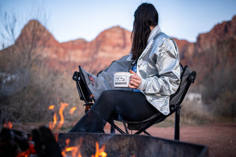 A woman sitting in a chair next to a campfire holding a coffee mug