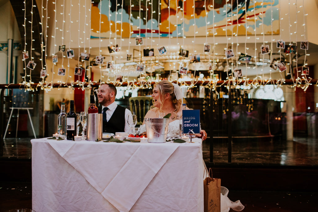 Bride and groom on sweetheart table at wedding breakfast with fairy lights and photo wedding stationery