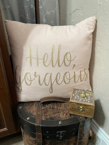 A pink pillow with Hello Gorgeous written on it in gold writing.  It is sitting on top of a treasure chest.
