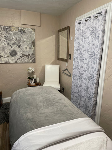 A cozy facial bed is beside a circular wooden table with a white orchid sitting on it.  In the corner, sits a comfortable white leather chair.  A beautiful gray and white floral curtain hangs in the entry way.
