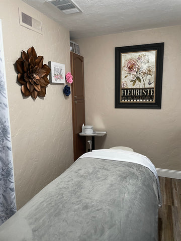 A serene treatment room with pink and copper floral artwork.  The facial bed has a gray soft and plush blanket for a relaxing and cozy experience.