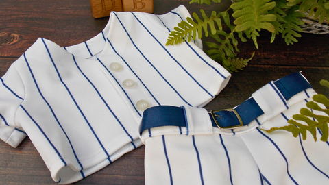 Teddy bear baseball uniform lying on a brown wooden surface. The teddy bear baseball uniform is made from a white fabric with a thin blue striped. It has a blue belt with a silver buckle. There is a green fern plan to the right of the picture