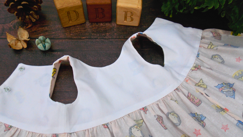 A teddy bear dress lying on a wooden surface with three wooden bricks in the top of the picture. The photo shows the inside of the teddy bear dress with a white lined top section.