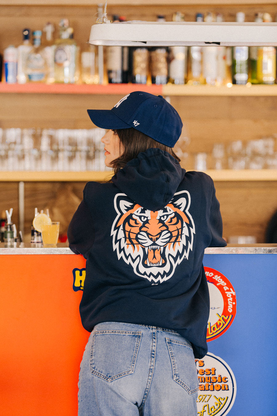 Mädchen an der Bar im Total-Look der Marke 47 mit Sweatshirt mit Tigergrafik auf der Rückseite und blauer, gebogener Schirmmütze der NY Yankees