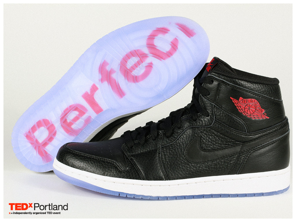 Pair of mid top sneakers, Air Jordan 1 "Perfect" x TEDx Portland, black colorway in textured leather, semi-transparent icy blue outer sole showing "Perfect" writing on the midsole