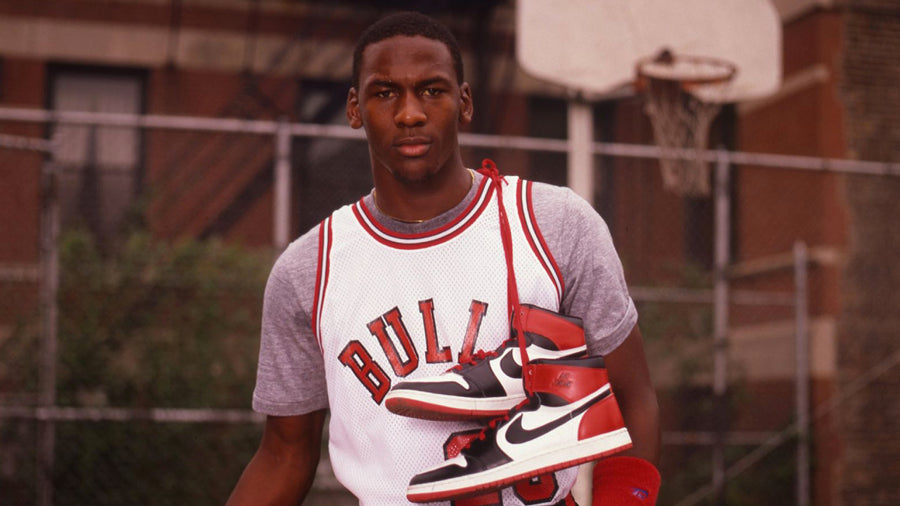Michael Jordan in an urban basketball playground wearing Chicago Bulls jersey number 23 with the Air Jordan 1 OG shoes in the white, red and black Banned colorway on his shoulders