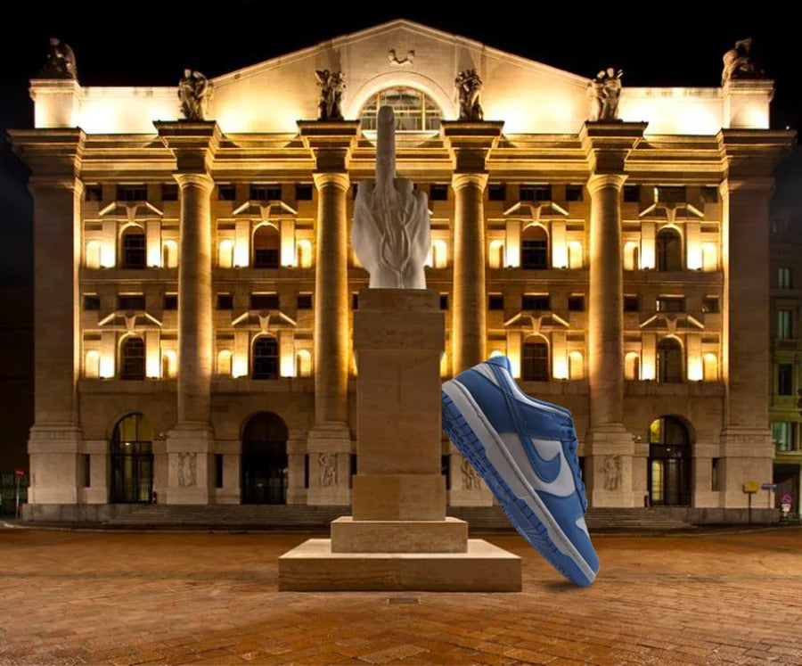 Sneaker gigante Nike Dunk Low Retro UNC appoggiata alla scultura del Dito Medio di Cattelan nella Piazza della Borsa di Milano