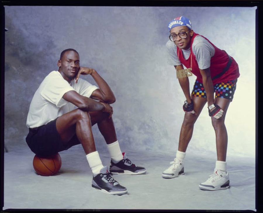 On the left Michael Jordan sitting on a basketball with Air Jordans on his feet and on the right Stan Lee with a sporty 90s look with a pair of white Air Jordan basketball sneakers on his feet