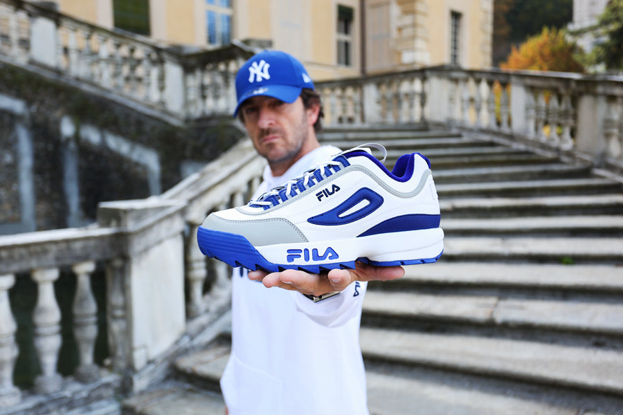 Man dressed in street style with a blue New York Yankees curved visor cap holding Fila x Atipici Disruptor "Blueprint" sneakers in the white blue colorway