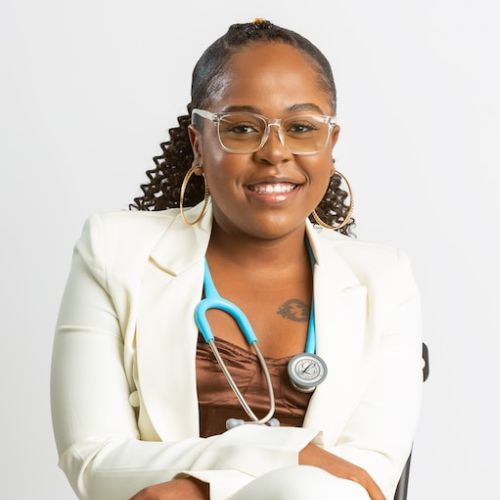 Doctoresse peau métisse avec une blouse blanche, des lunettes et un stéthoscope bleue assise devant un fond blanc