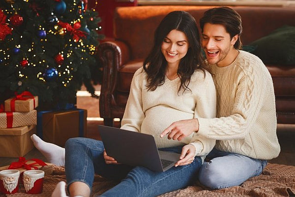 4 pregnant couple sat by a Christmas tree looking at a laptop