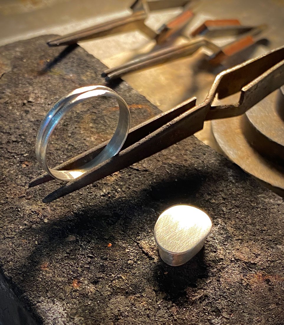 A ring in metal forceps and the polished silver setting beside it await the fire that will combine them into a single piece of jewellery.