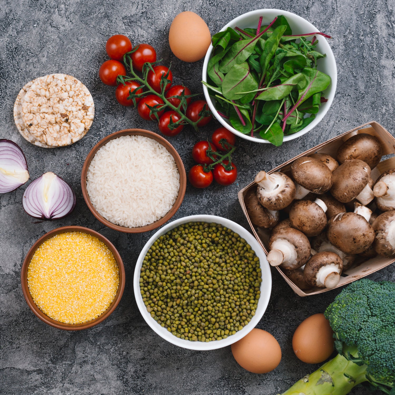 Bowls of rice grains; mung beans; polenta with fresh vegetables; eggs and puffed rice cake on gray textured backdrop