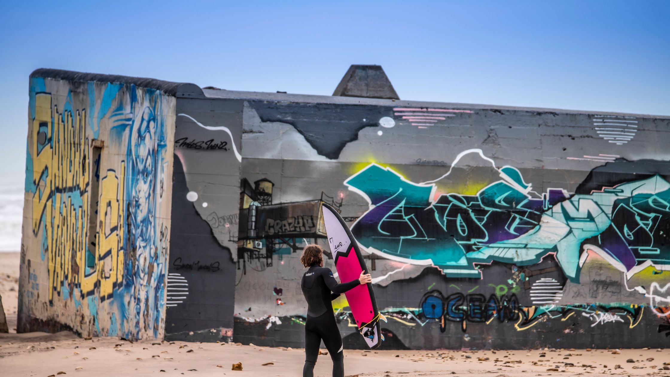 A surfer in front of a blockhouse with a Zeus surfboard
