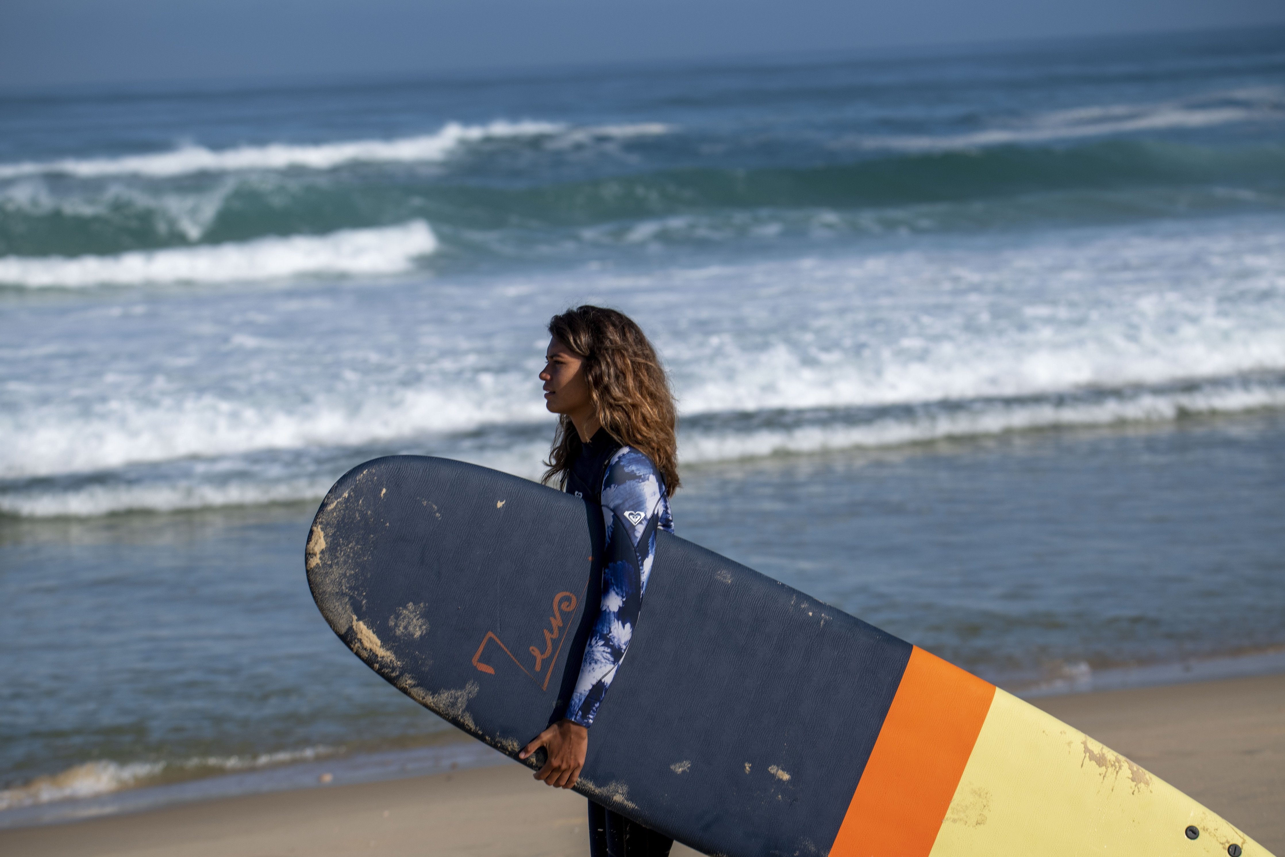 Surfista femenina llevando una tabla de surf XXL Zeus azul marino, naranja y amarillo en la playa