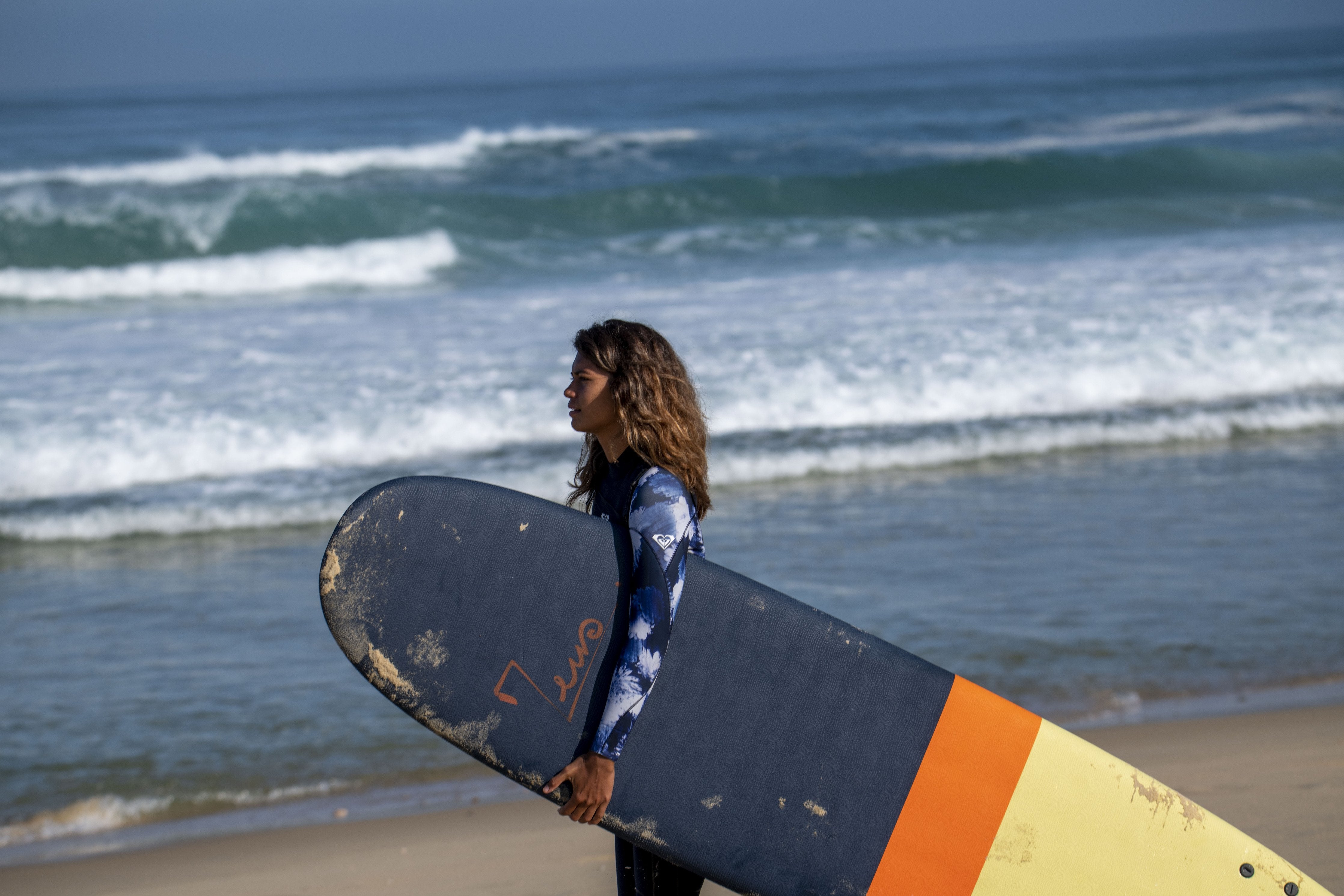 Surfeuse portant une planche de Surf XXL Zeus Bleu marine orange et jaune sur la plage