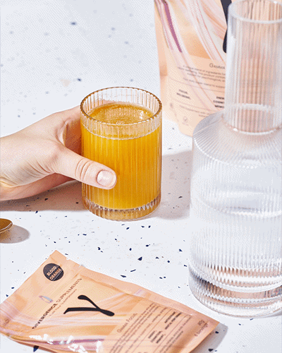 A hand holding a ribbed glass with orange liquid, next to beauty products and a clear ribbed bottle.