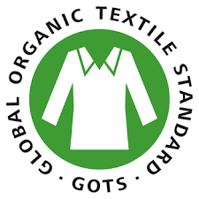 Il s'agit d'un logo circulaire avec un fond vert et une bordure blanche. Au centre, il y a un symbole blanc qui ressemble à une montagne avec deux sommets et un soleil levant derrière. Autour du cercle, il y a du texte en majuscules qui dit "GLOBAL ORGANIC TEXTILE STANDARD" avec les mots "GLOBAL" et "STANDARD" en haut et "ORGANIC TEXTILE" en bas. En dessous du cercle, il y a l'acronyme "GOTS" en lettres majuscules.