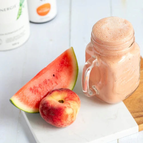Cut watermelon and a peach next to a jam jar filled with light pink watermelon and peach smoothie with XbyX Daily Energy and XbyX Take It Easy products in the background