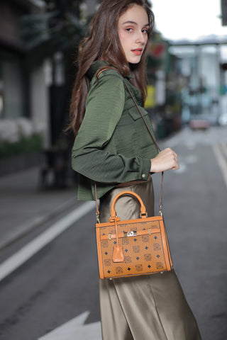 A young woman looks over her shoulder carrying a brown Saga handbag, a green blouse and beige pants on the street.