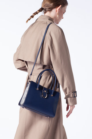 A woman stands with her back holding a dark blue leather women's handbag with decorative details in her hand and wearing a beige coat.