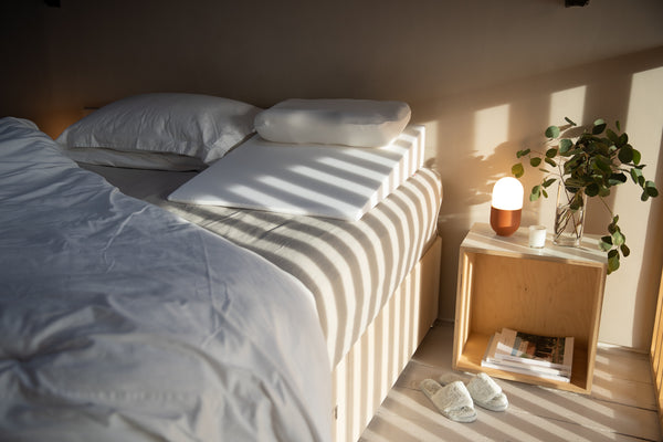 an empty bed with white bed sheets and Putnams duvet and a wooden bedside table with lamp and plant on it