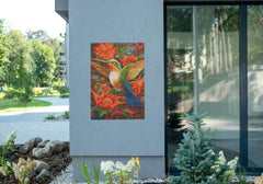 Diamond painting van een kolibrie tussen oranje bloemen opgehangen aan een grijze muur buitenshuis.