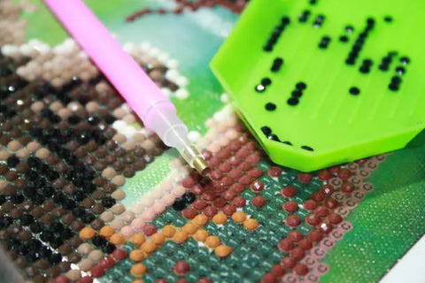 Detail van diamond painting met roze pen en groen bakje vol zwarte steentjes op kleurrijk patroon