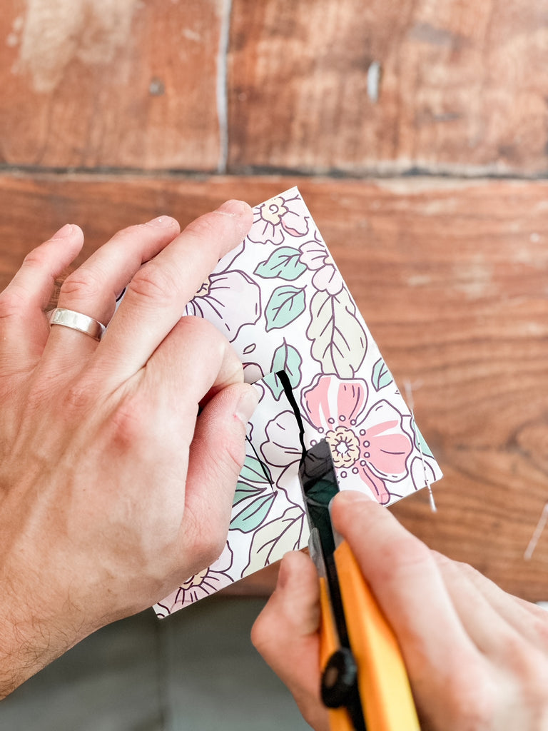 The image shows hands cutting a floral-patterned paper with a utility knife on a wooden surface, indicative of a craft activity.