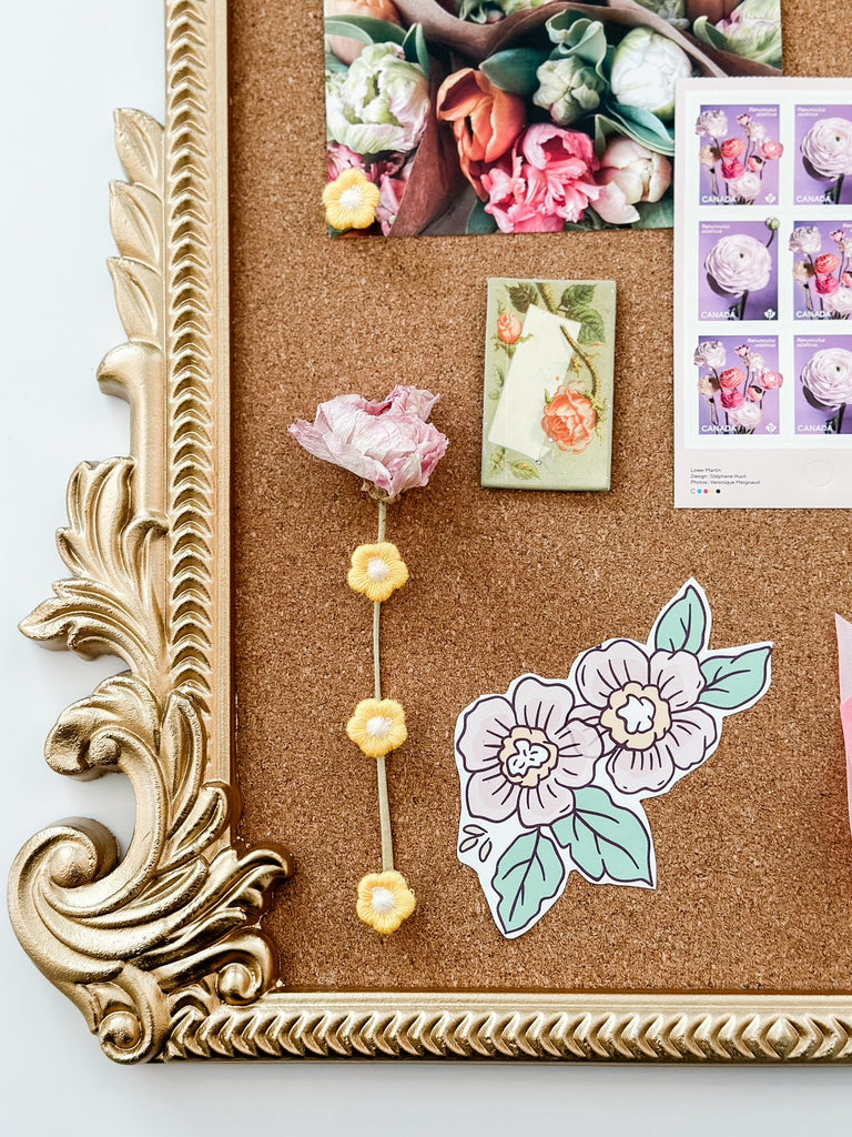 A close-up view of a portion of a corkboard with a gold ornate frame. On the board, there's a card with a floral design, a few pastel-colored flower illustrations, and a three-dimensional paper flower attached with a yellow pom-pom string.