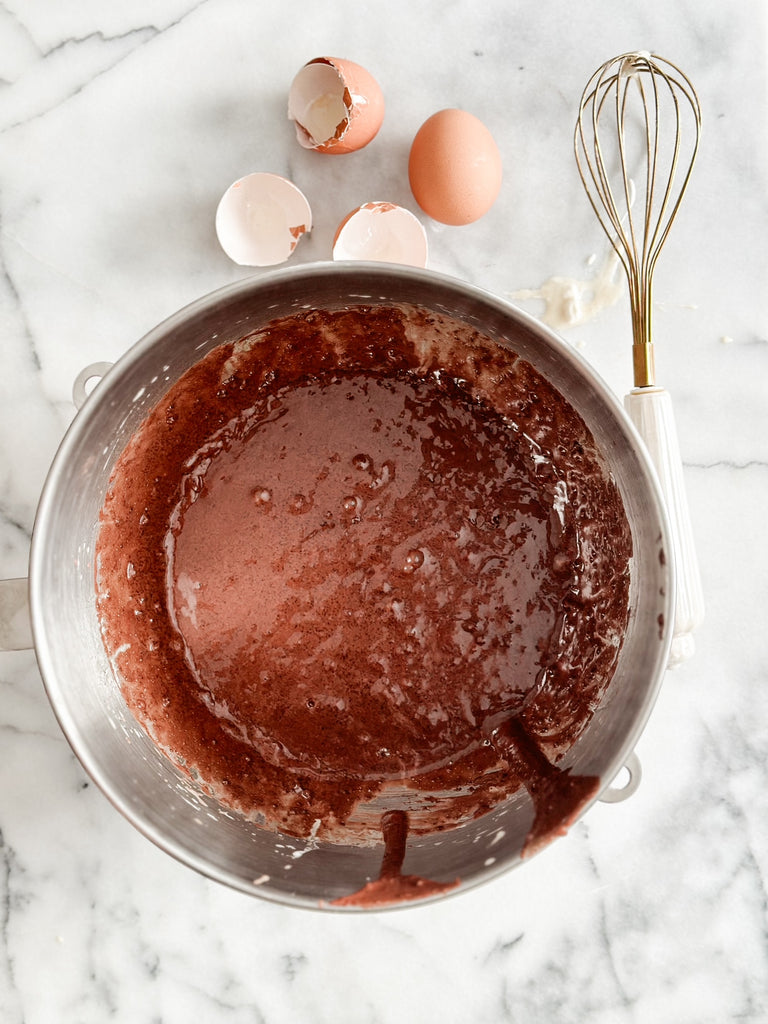 A stainless steel mixing bowl filled with chocolate batter, a whisk, and eggshells on the side, placed on a marble surface.