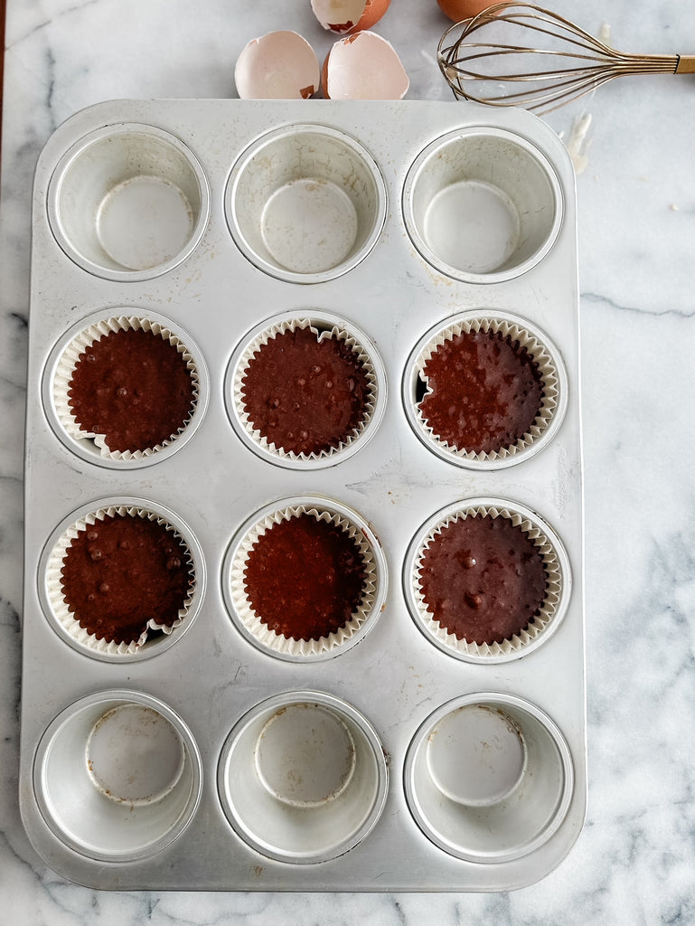 A muffin tin with six chocolate cupcake batters in white liners and empty greased cups, on a marble countertop with a whisk, eggshells, and a measuring cup visible.