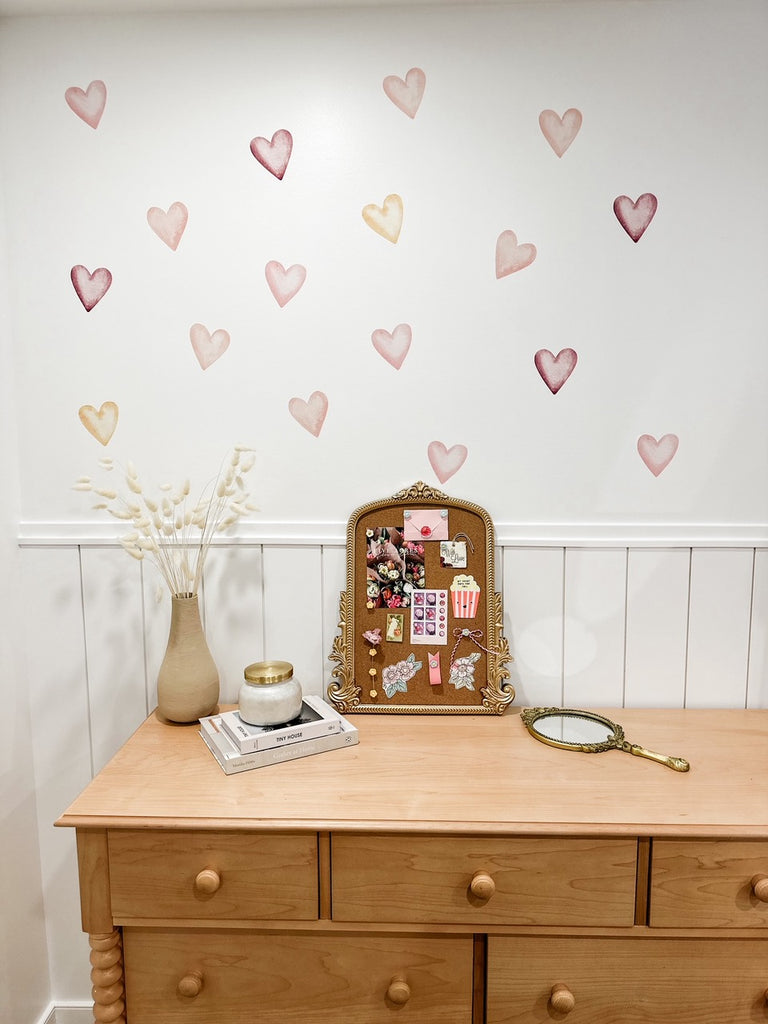 A wider room view where the corkboard is placed on a wooden dresser against a wall with heart-shaped decals in various shades of pink. The room's decor includes a vase with white botanicals, books, and a magnifying glass, creating a charming and cozy Valentine's Day themed space.