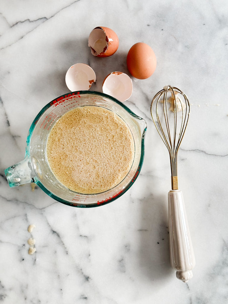 A measuring cup filled with a frothy, bubbly egg white mixture next to eggshells and a whisk, on a marble countertop