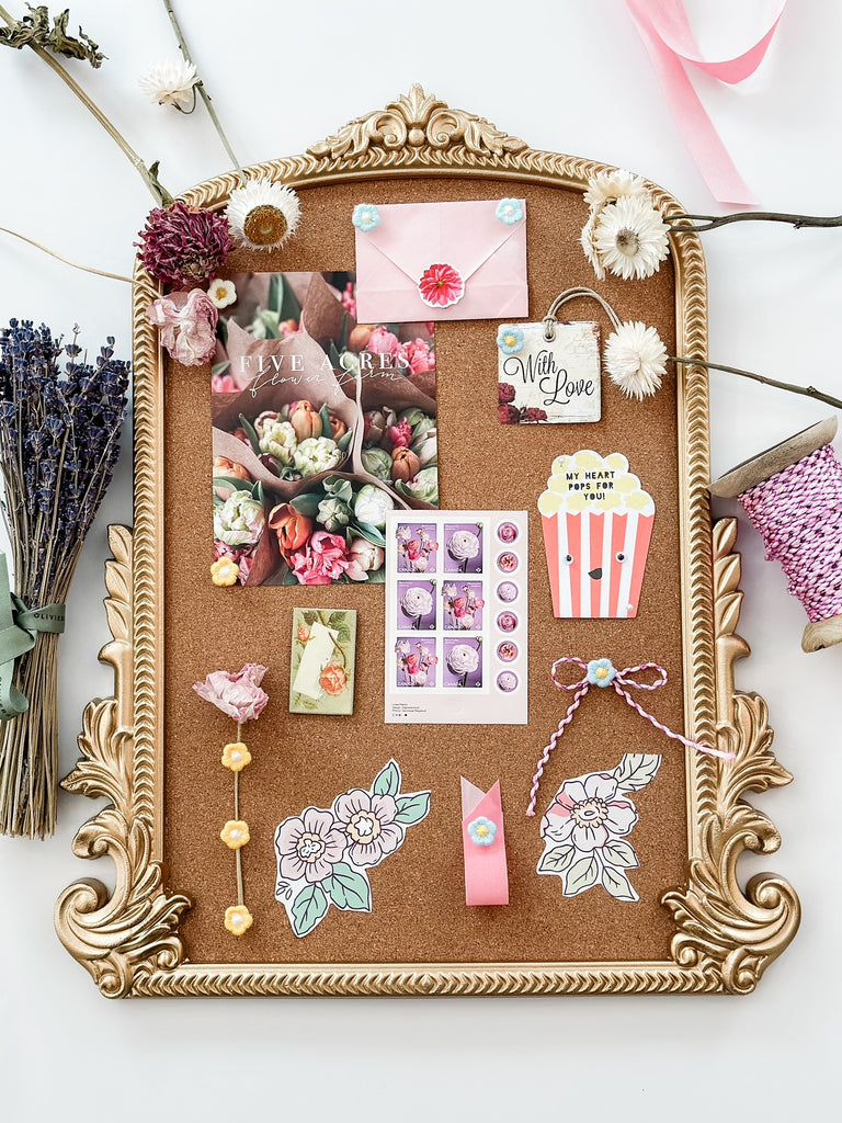 A full view of the same corkboard, showcasing an eclectic assortment of items pinned to it, including floral prints, a 'With Love' tag, a popcorn-shaped card, and lavender sprigs. The board is embellished with a pink ribbon bow, a blue floral envelope, and a string of decorative hearts, all set against a soft-hued background.