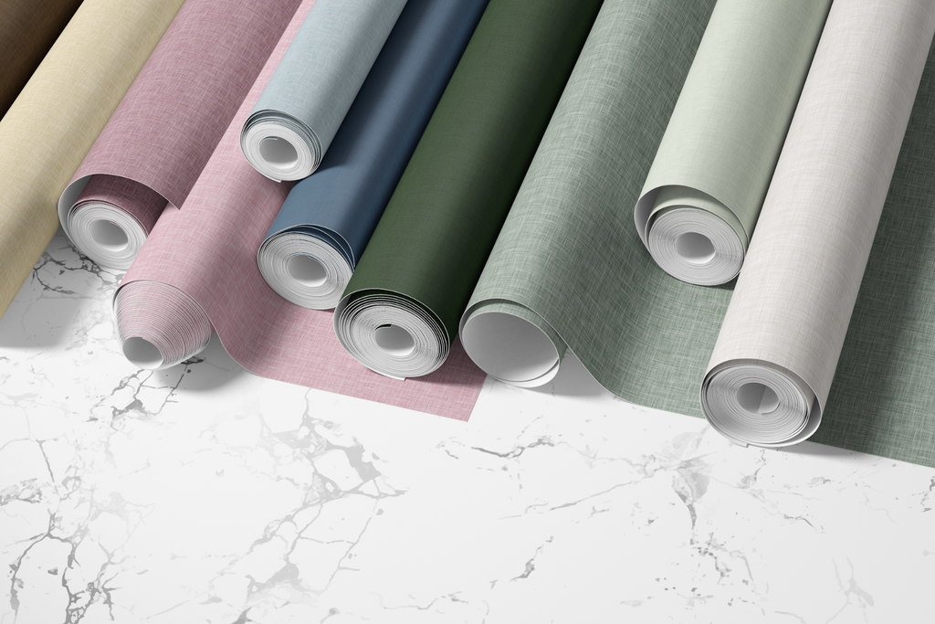 An array of peel and stick wallpapers with a grasscloth texture, displayed in various colors. From left to right, the rolls are cream, light pink, sky blue, navy blue, forest green, sage green, and light gray. Each roll is partially unrolled, showing the grasscloth texture and color