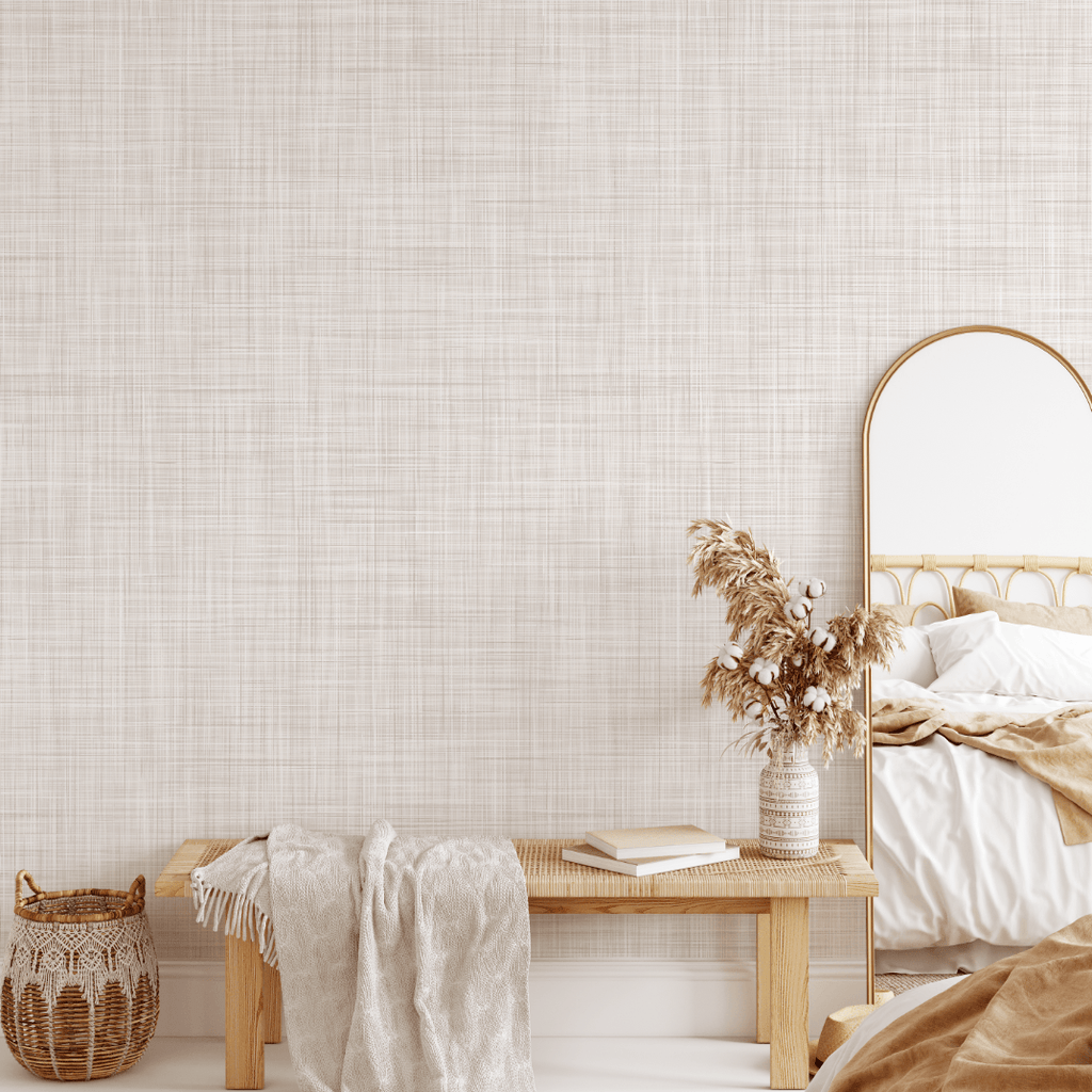 A cozy bedroom corner showcasing a wall covered in peel and stick wallpaper with a subtle seagrass linen texture. The room features a natural wood bench with a cream-colored throw and a couple of books on top, beside a wicker basket.