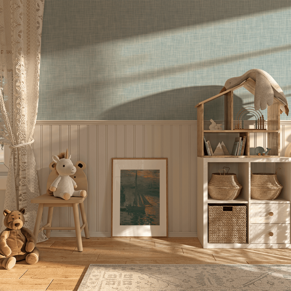 A children's playroom corner with sky-blue grasscloth peel and stick wallpaper, creating a serene backdrop. The room is bathed in warm sunlight filtering through a lace curtain, highlighting a white beadboard and the soft textures of the wallpaper. A stuffed bear sits on the wooden floor, beside a stool with a plush toy horse and under a shelf with baskets, books, and a fabric elephant.