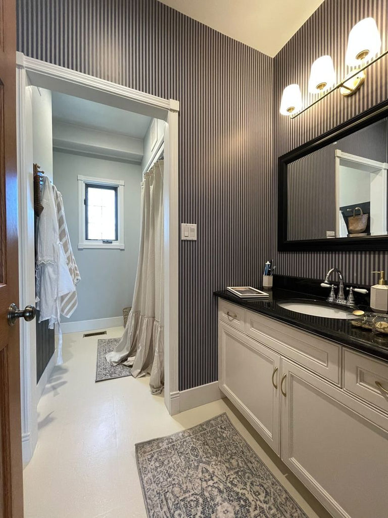 A country club bathroom with sleek striped wallpaper in shades of gray. White trim and cabinetry provide a crisp contrast, while a classic patterned area rug adds texture. A well-lit vanity mirror and soft window treatments create a bright and elegant space.