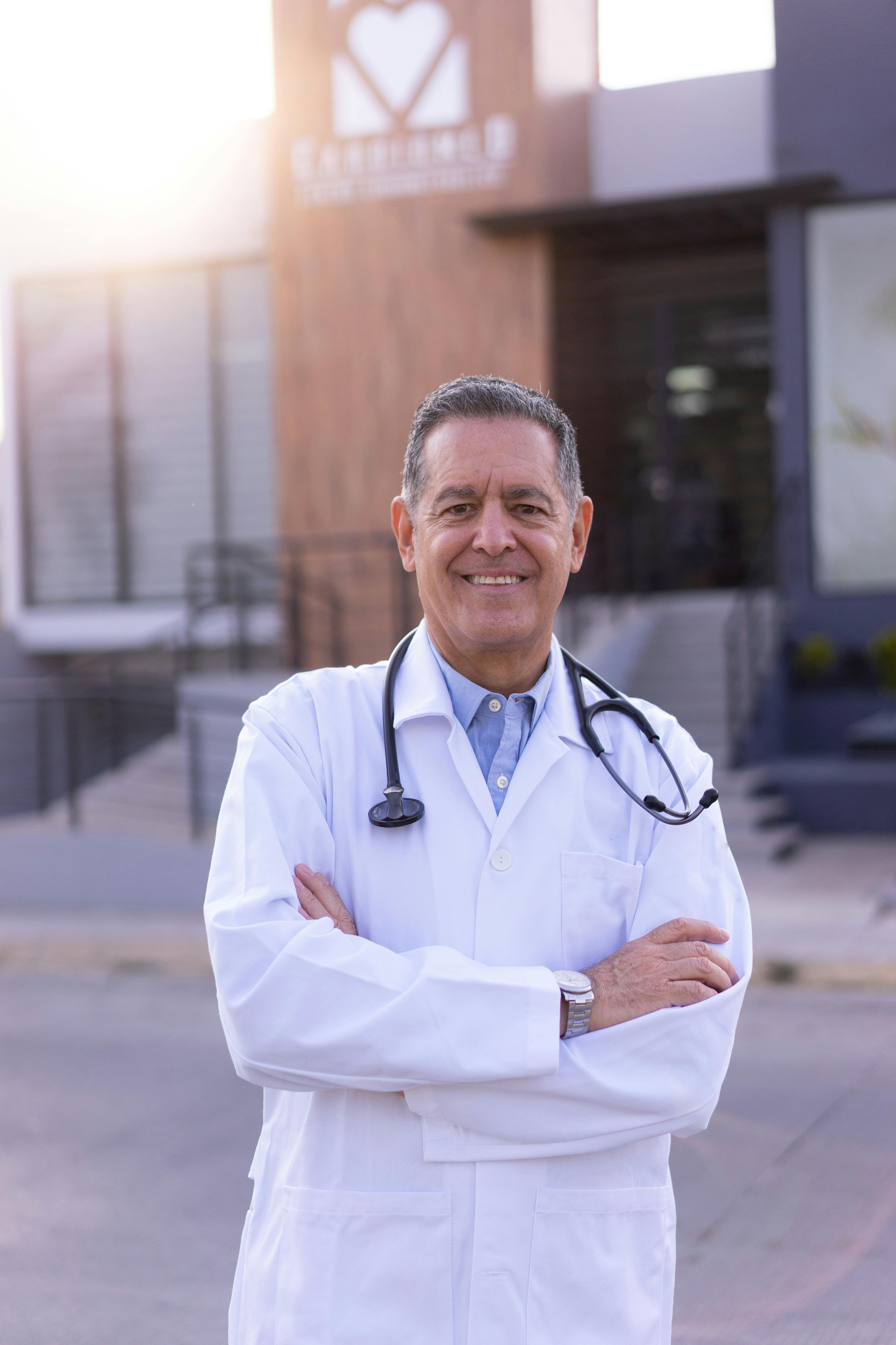 Smiling male doctor with stethoscope standing in front of a clinic at sunset.