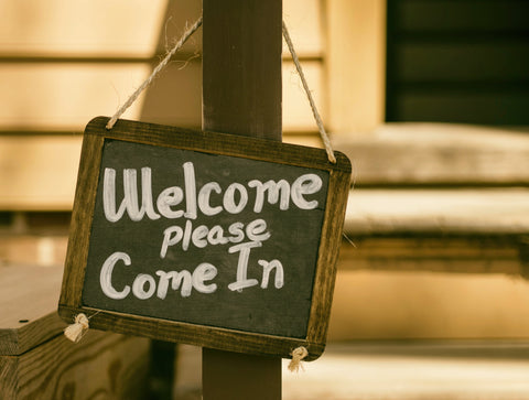 A rustic sign hanging on a string. Text: Welcome, Please Come In