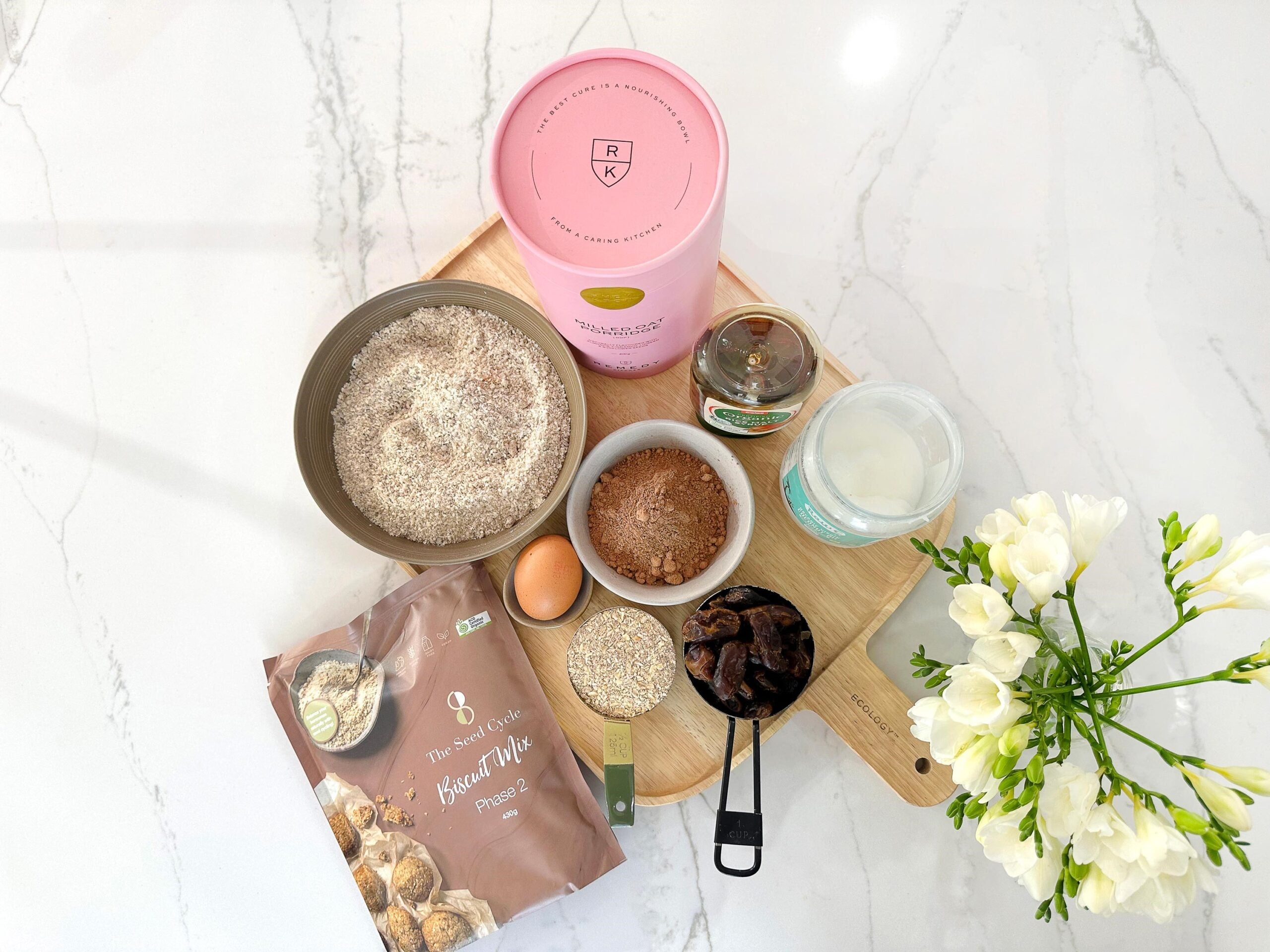 flat lay of seed cycle biscuit mix package, dry mix in bowl, chopping board, flowers and measuring cup.