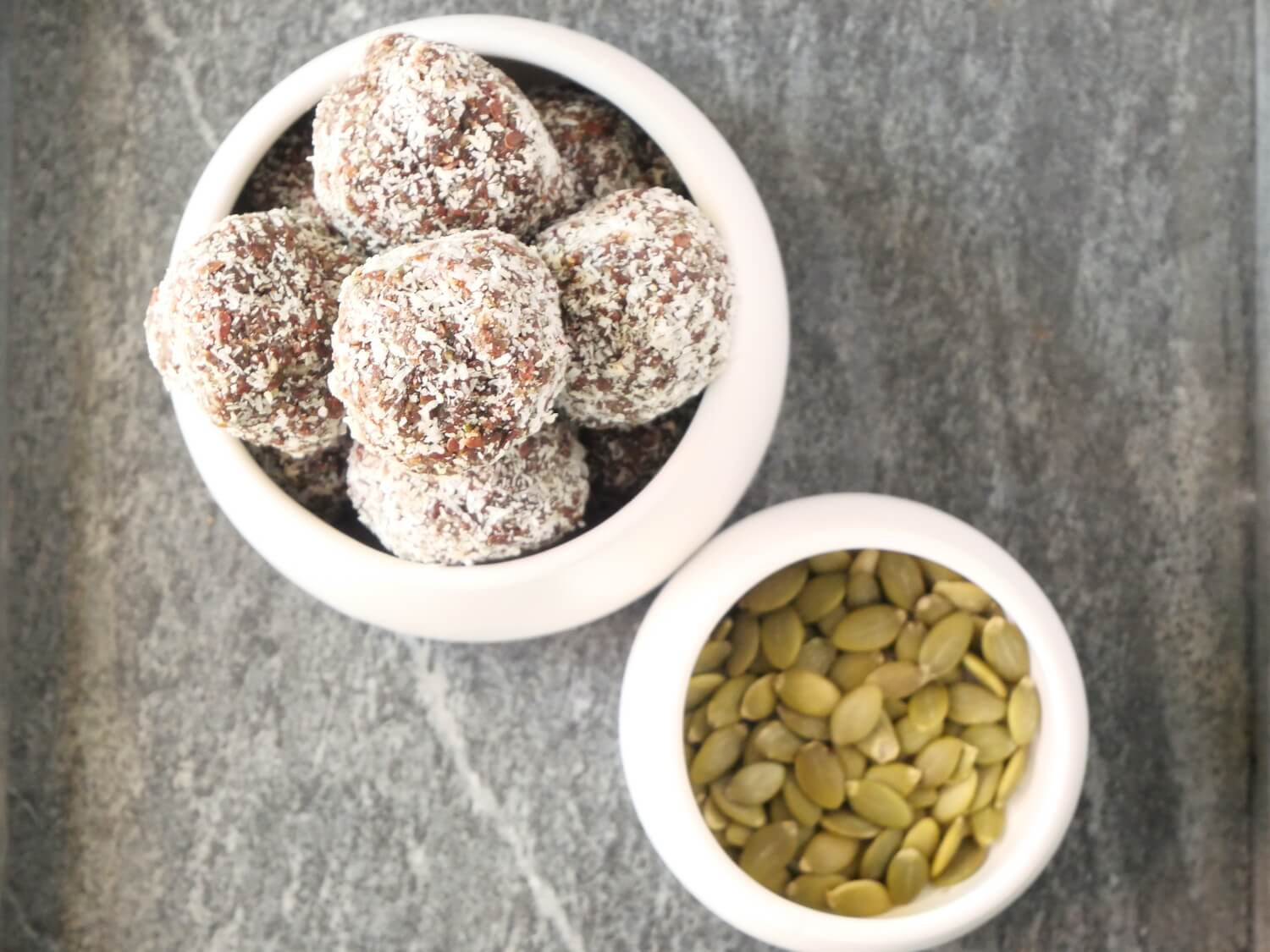 Two white bowls on a kitchen bench. The first bowl is full of chocolate seed cycle bliss balls. The second bowl is filled with pumpkin seeds.