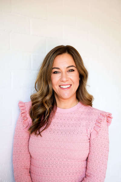 Melissa Kovacevic, the founder of The Seed Cycle, standing in front of a white brick wall, wearing a pink knitted jumper with decorative shoulders. About The Seed Cycle.