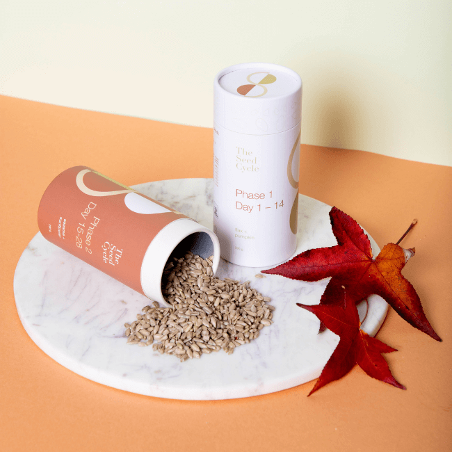 Two canisters 'of The Seed Cycle product on a circular marble tray. One of the canisters is laying on it's side with seed pouring out. There are two Autumn leaves sitting next to the packaging, which is on an orange and creamy yellow backdrop. The Seed Cycle Subscription.