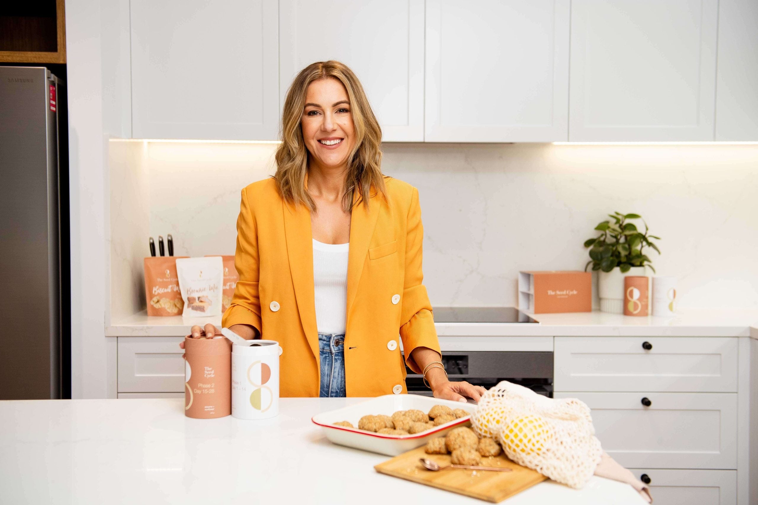 Melissa Kovacevic founder of The Seed Cycle in white kitchen with products wearing jeans and orange blazer