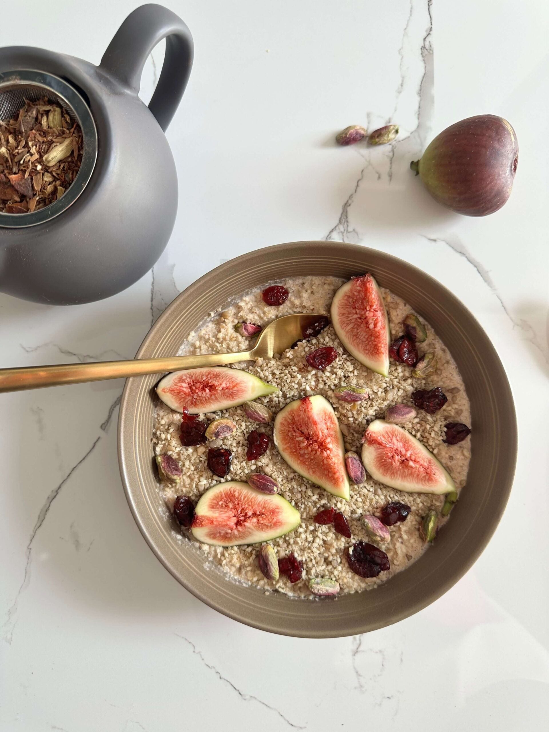 Seed + Porridge with Fig, Cranberry, and Pistachios with gold spoon and grey tea cup with tea leaves visable