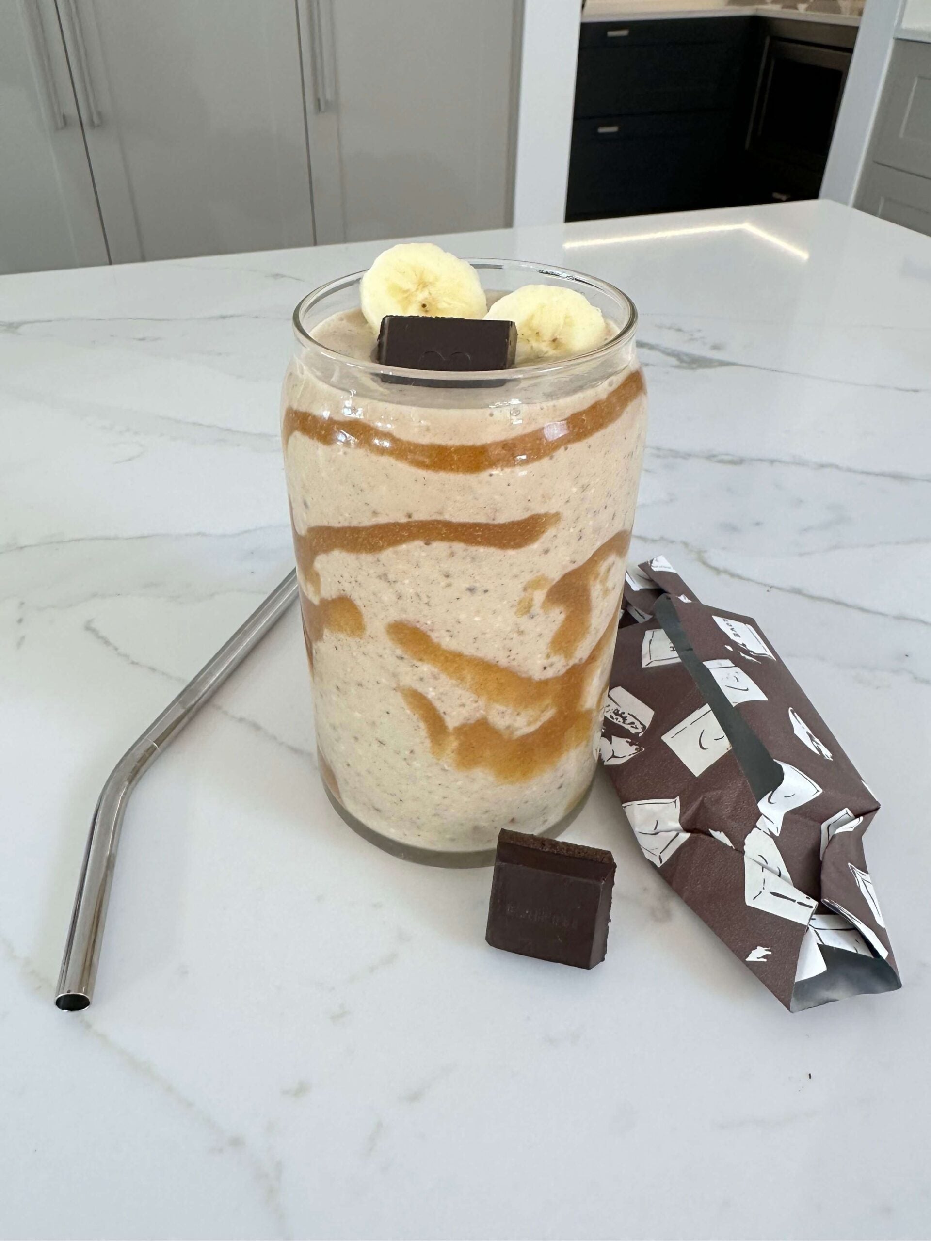 smoothie glass with date and banana smoothie topped with a square of chocolate and sliced banana on kitchen bench with straw placed next to the glass.